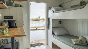 Een kleine kamer met stapelbedden, een houten tafel en uitzicht op een waterpartij door een geopende deur.