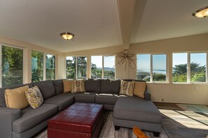 A spacious living room with a large sectional sofa, a red ottoman, and large windows offering a view of the outdoors. - Moonstone Beach Retreat - Beach Lover's Getaway! (Trinidad)