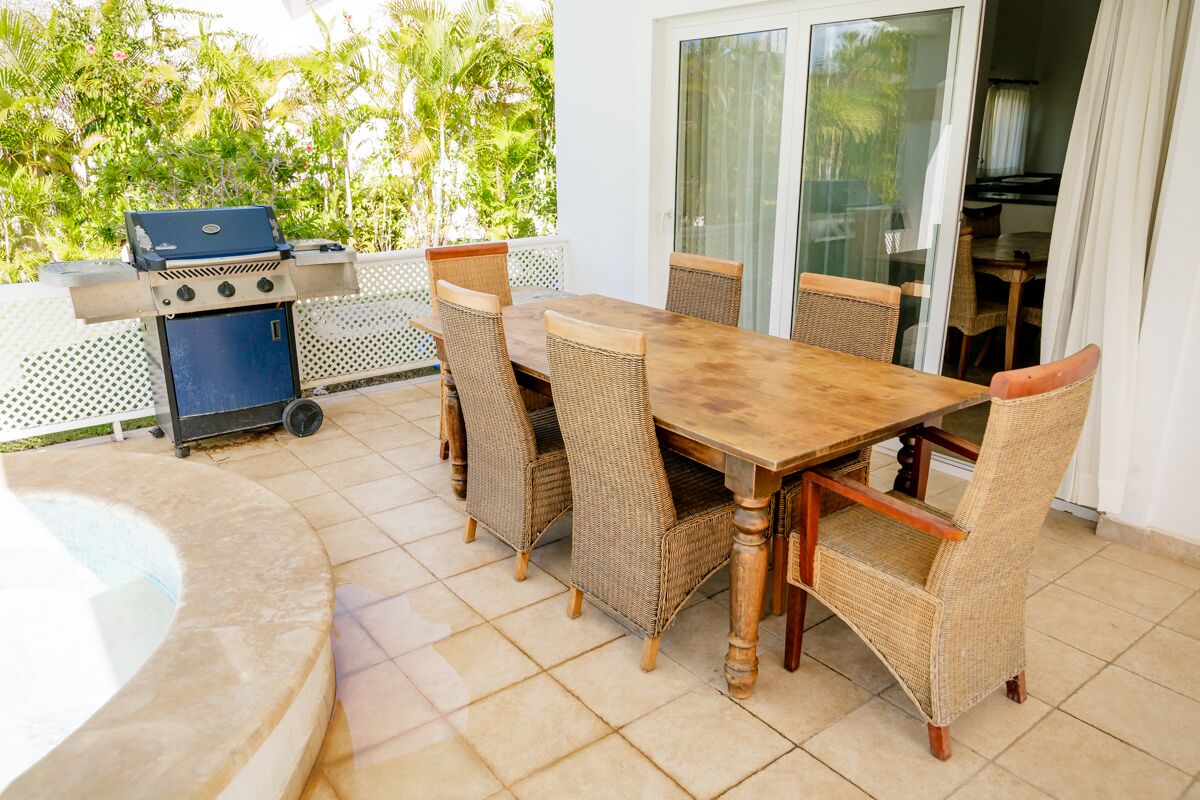 Een terras buiten met een houten tafel, rieten stoelen, een barbecue en een jacuzzi.