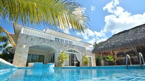 A pool area with a clear blue pool, a stone building with a white balcony, and a thatched-roof structure.