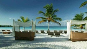 A beach with two wicker lounge chairs under white canopies, a palm tree, and a clear blue sky.