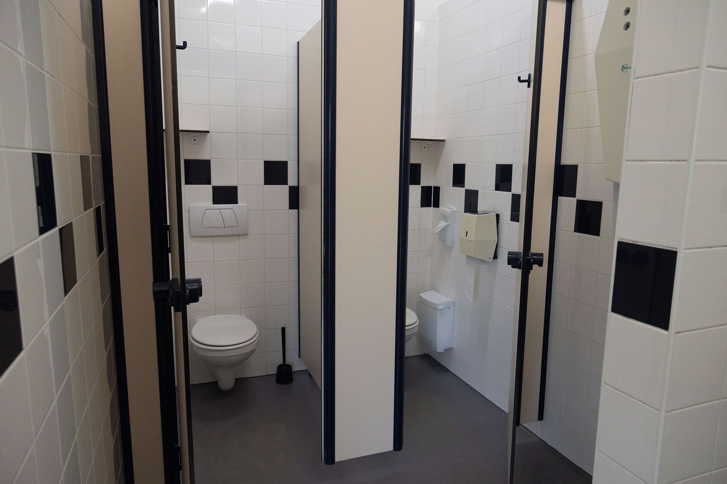 A restroom with white tiled walls, black and white checkered tiles, a white toilet, a black floor scrubber, and a hand dryer.
