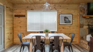 A wooden-paneled room with a dining table and chairs, a window with blinds, a hanging light fixture, and a stone fireplace.