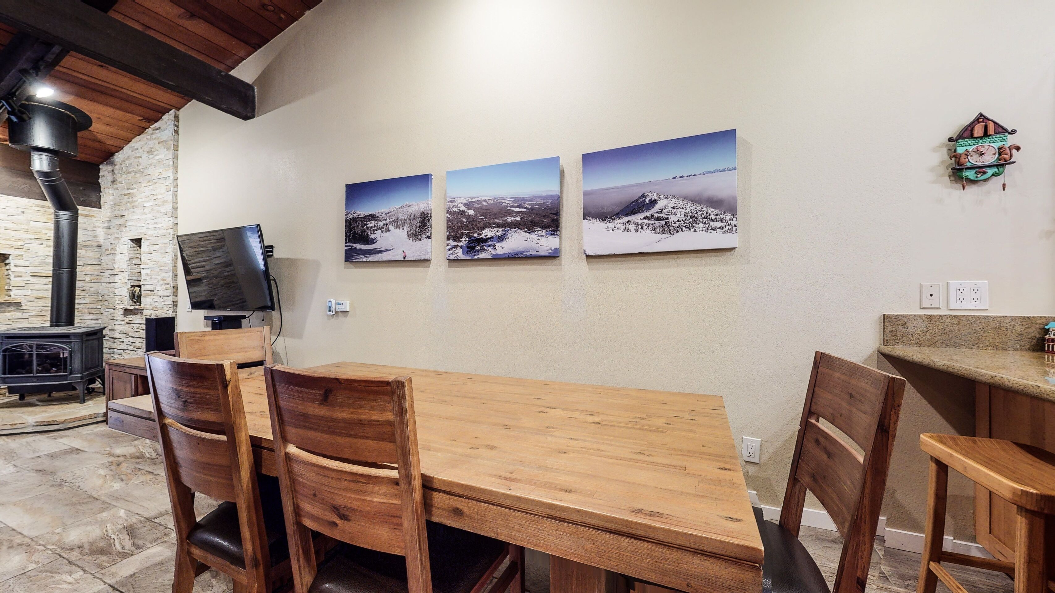 A dining area with a wooden table and chairs, a stone fireplace, and three framed landscape photographs on the wall.