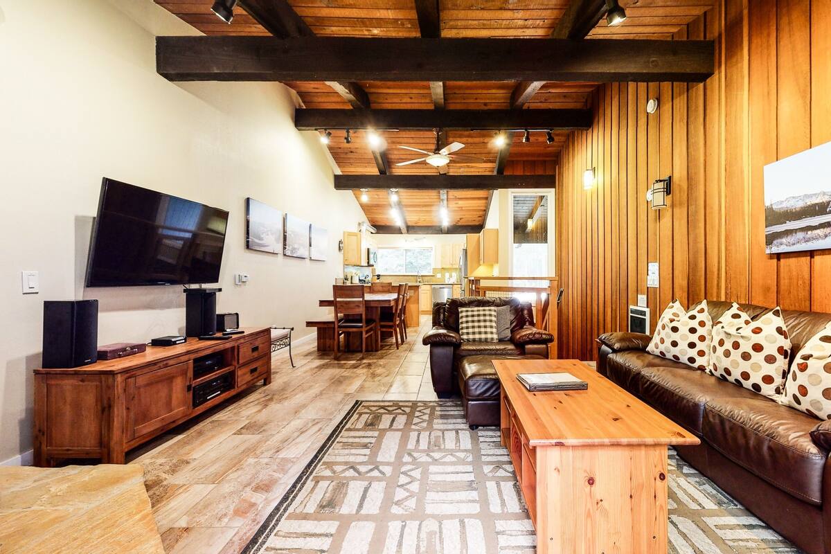 A cozy living room with a wooden ceiling, a leather sofa, a wooden coffee table, and a flat-screen TV on a wooden stand.