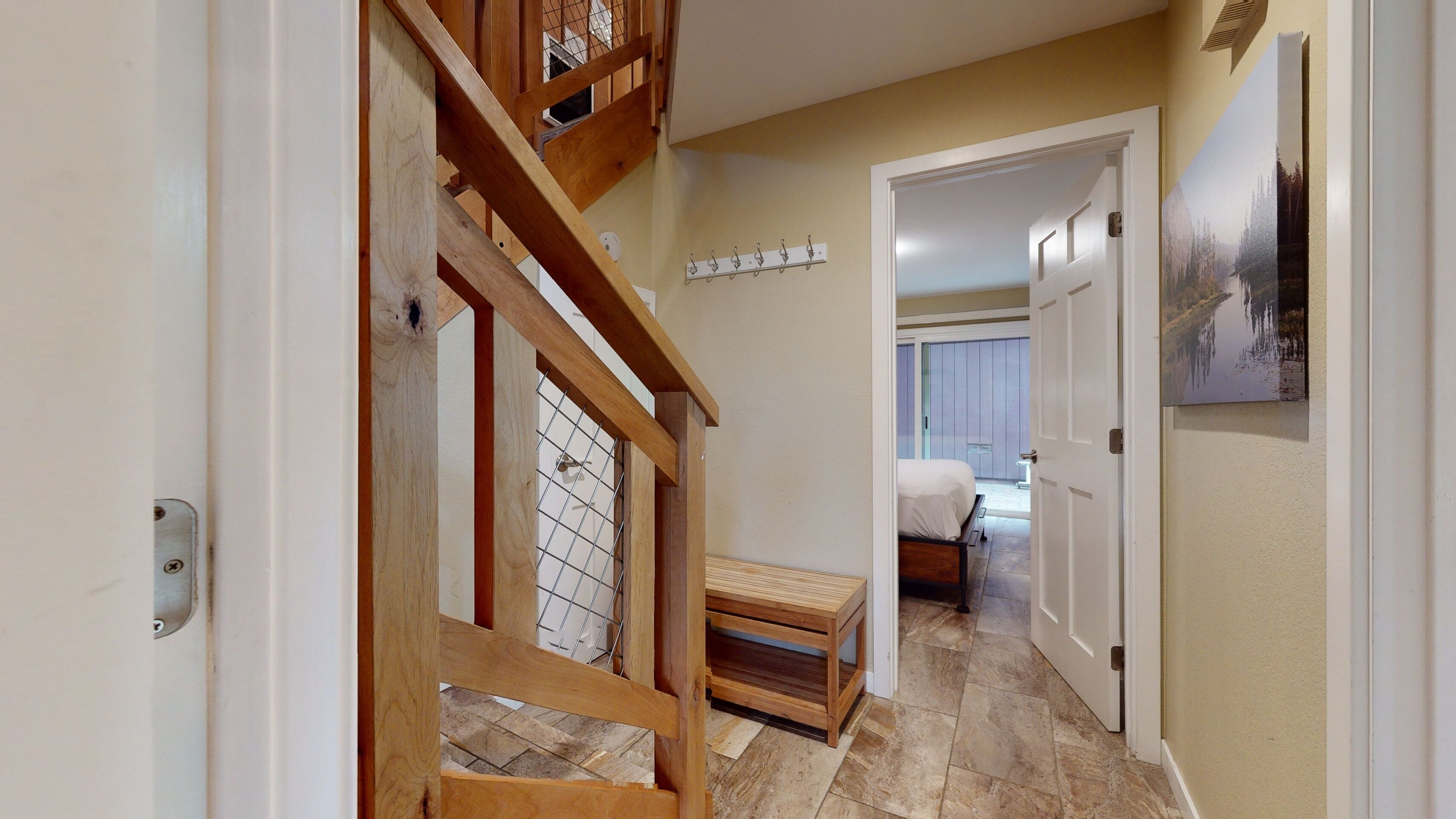 A hallway with a wooden staircase, a small bench, a bed in the adjacent room, and a door leading to another room.