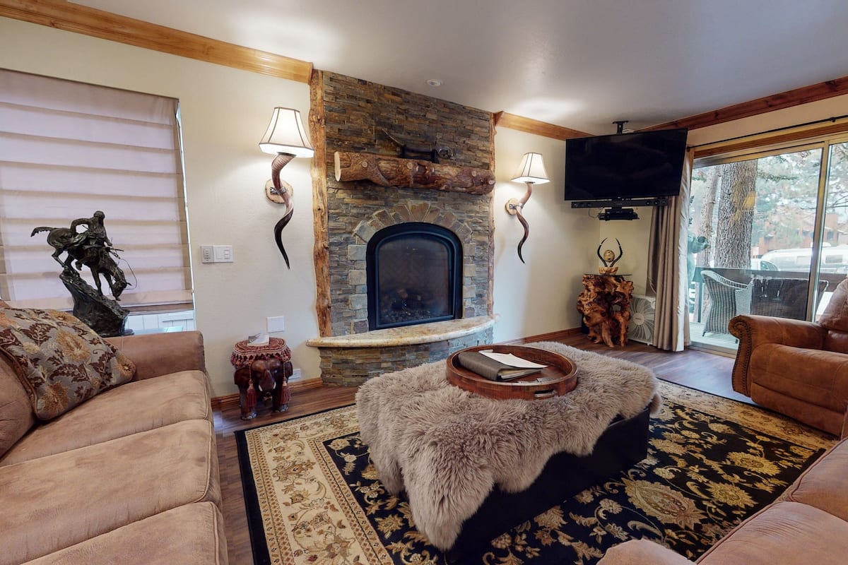 A cozy living room with a stone fireplace, leather sofas, and a large window with a view of trees.