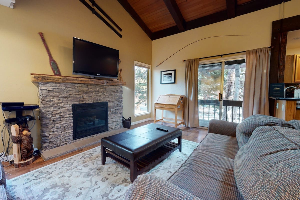 A cozy living room with a stone fireplace, a flat-screen TV mounted above it, a large sofa, and a dining area visible through a sliding glass door.