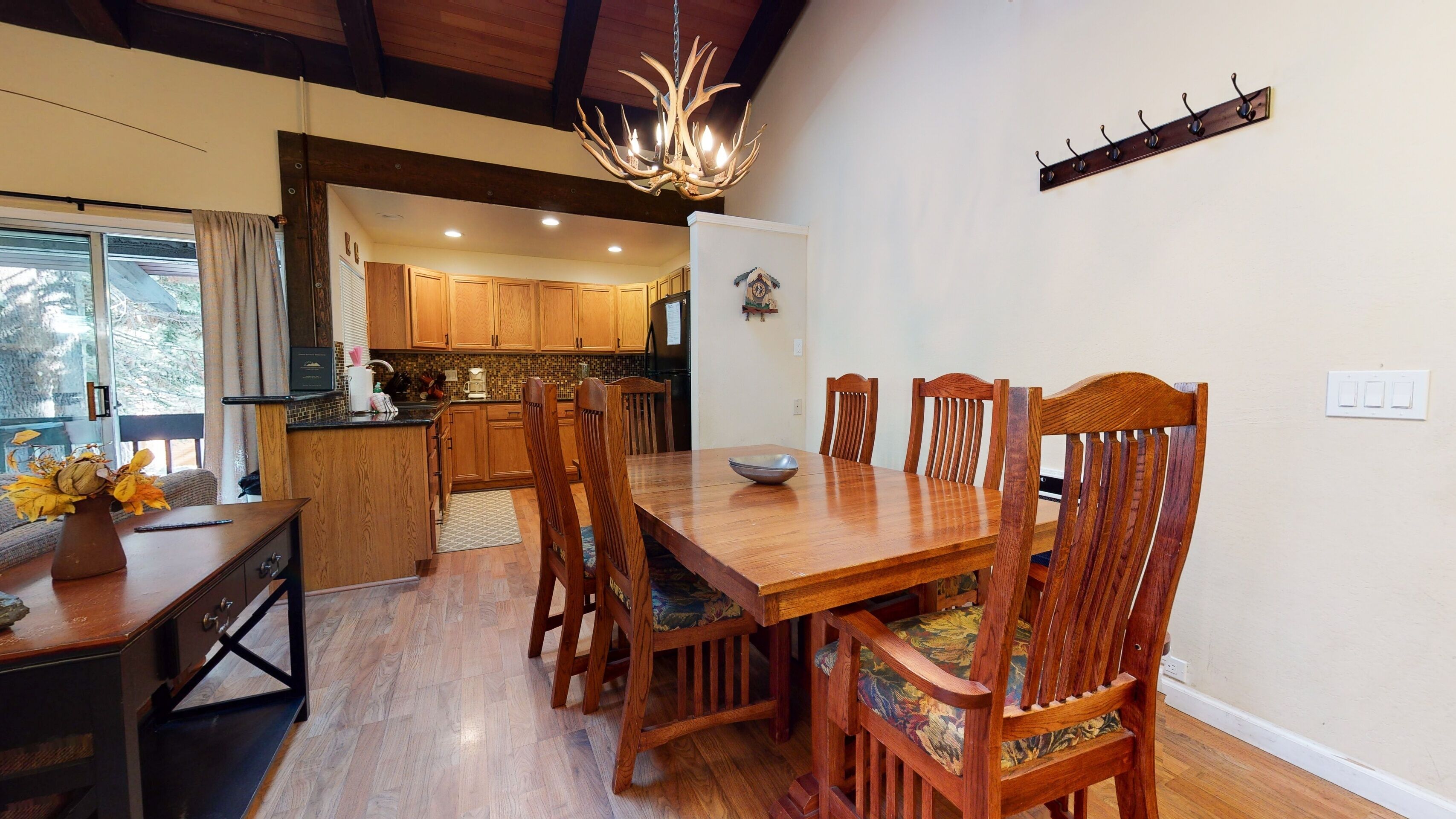 A dining area with a wooden table and chairs, a chandelier, and a kitchen in the background.