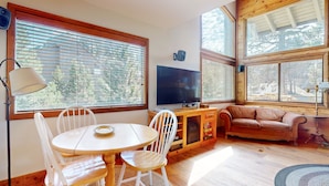 A cozy living room with a wooden table, white chairs, a brown sofa, and a television. Large windows with blinds allow natural light and views of the outdoors. - Rainbow Villas 1 Charming Boutique complex, Dual Master Suites, Forest Mountain Views by RedAwning (Mammoth Lakes)