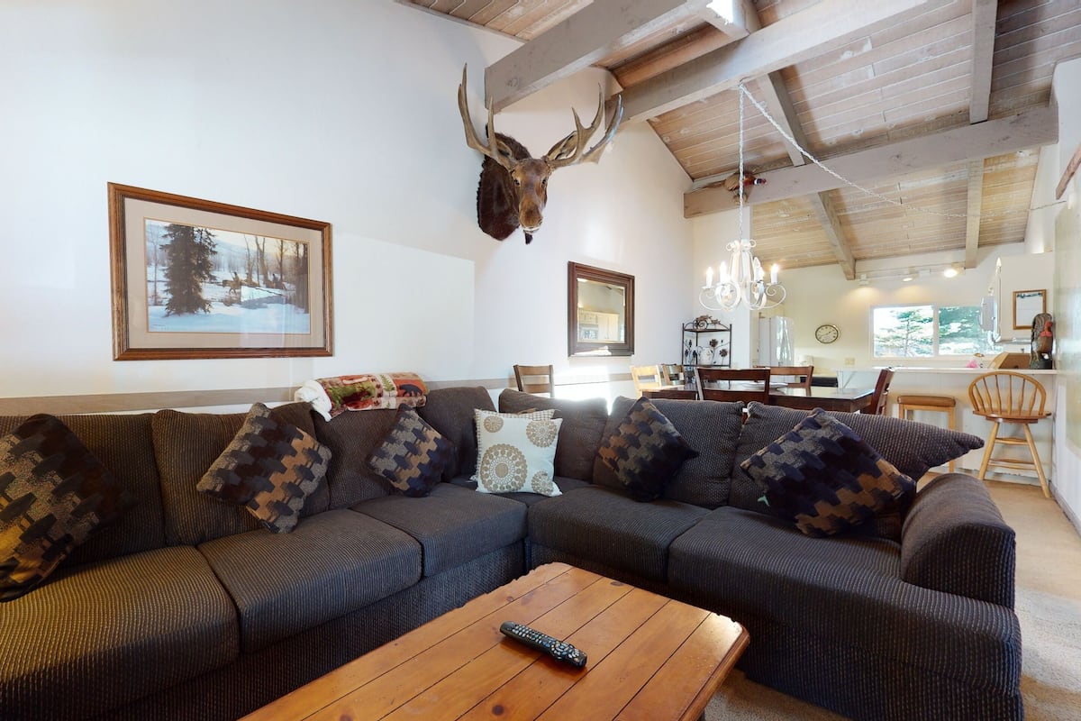 Ein Wohnzimmer mit Ecksofa, einem aufgehängten Hirschgeweih und einem Bilderrahmen mit einer verschneiten Winterlandschaft.