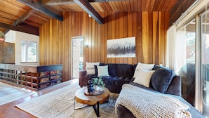 A cozy living room with a dark grey sofa, a round wooden coffee table, and a knitted throw. A large window allows natural light to fill the space.