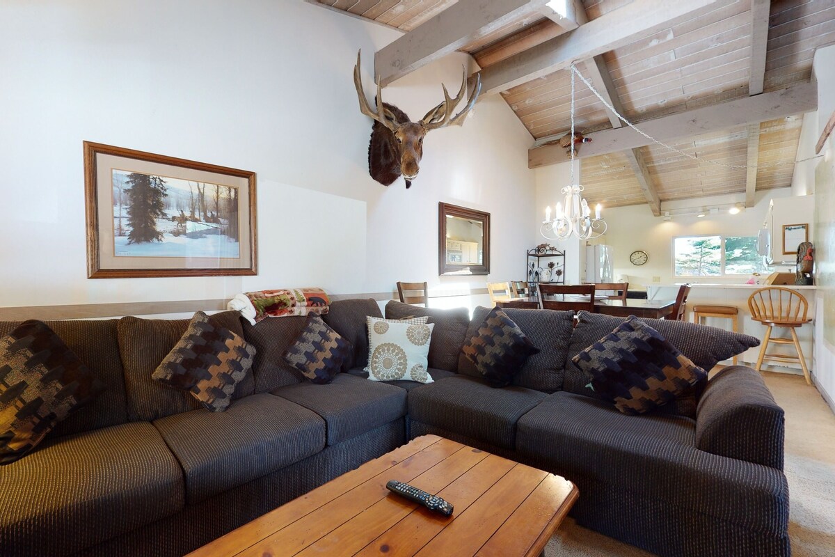 A living room with a sectional sofa, a mounted deer head, and a framed picture of a snowy landscape.