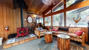 A cozy room with a red stove, wooden walls, a sofa with leaf patterns, and a large window with a deer head decoration.