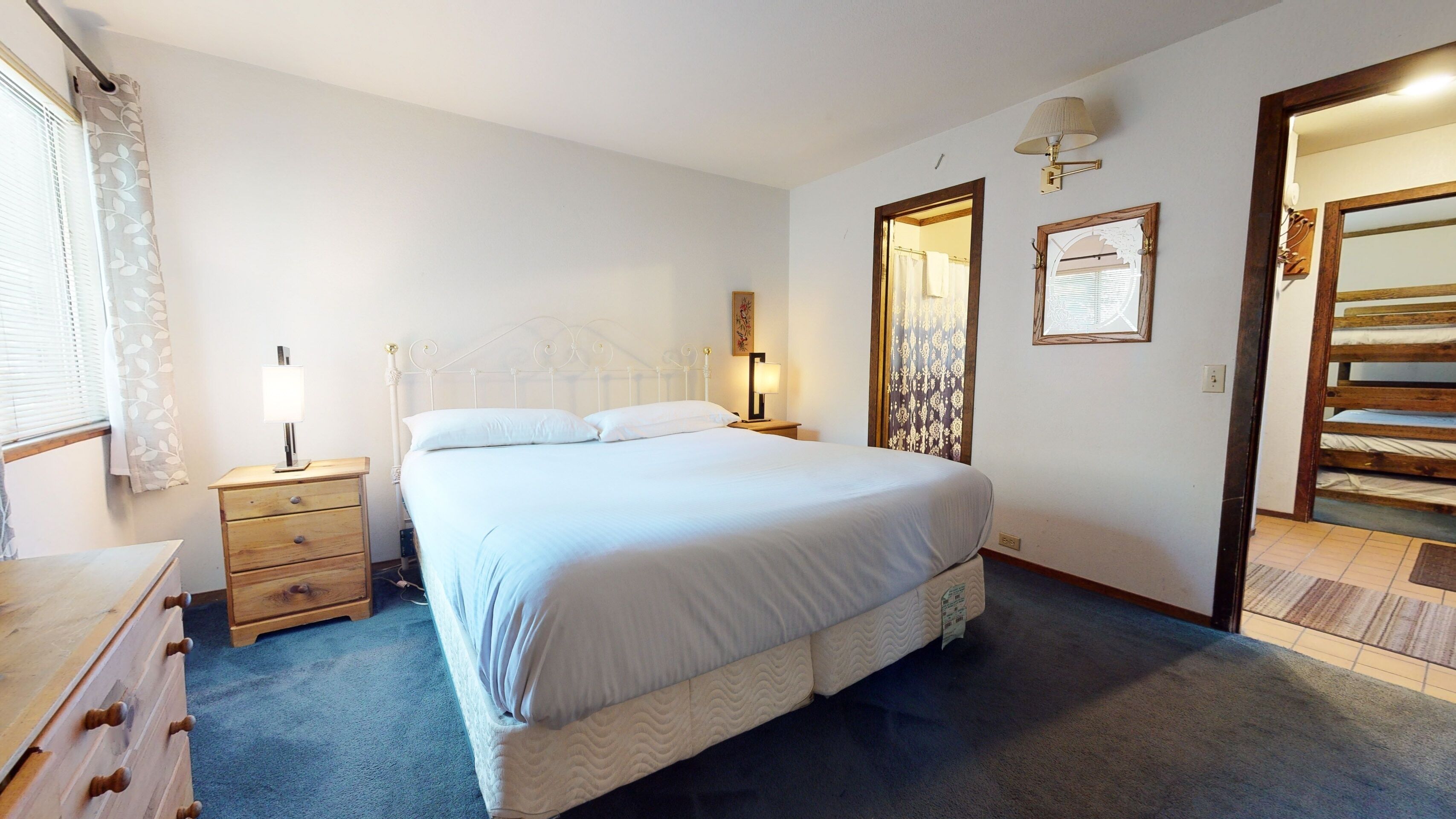 A bedroom with a bed, a nightstand, a dresser, a wall-mounted lamp, and a door leading to another room.