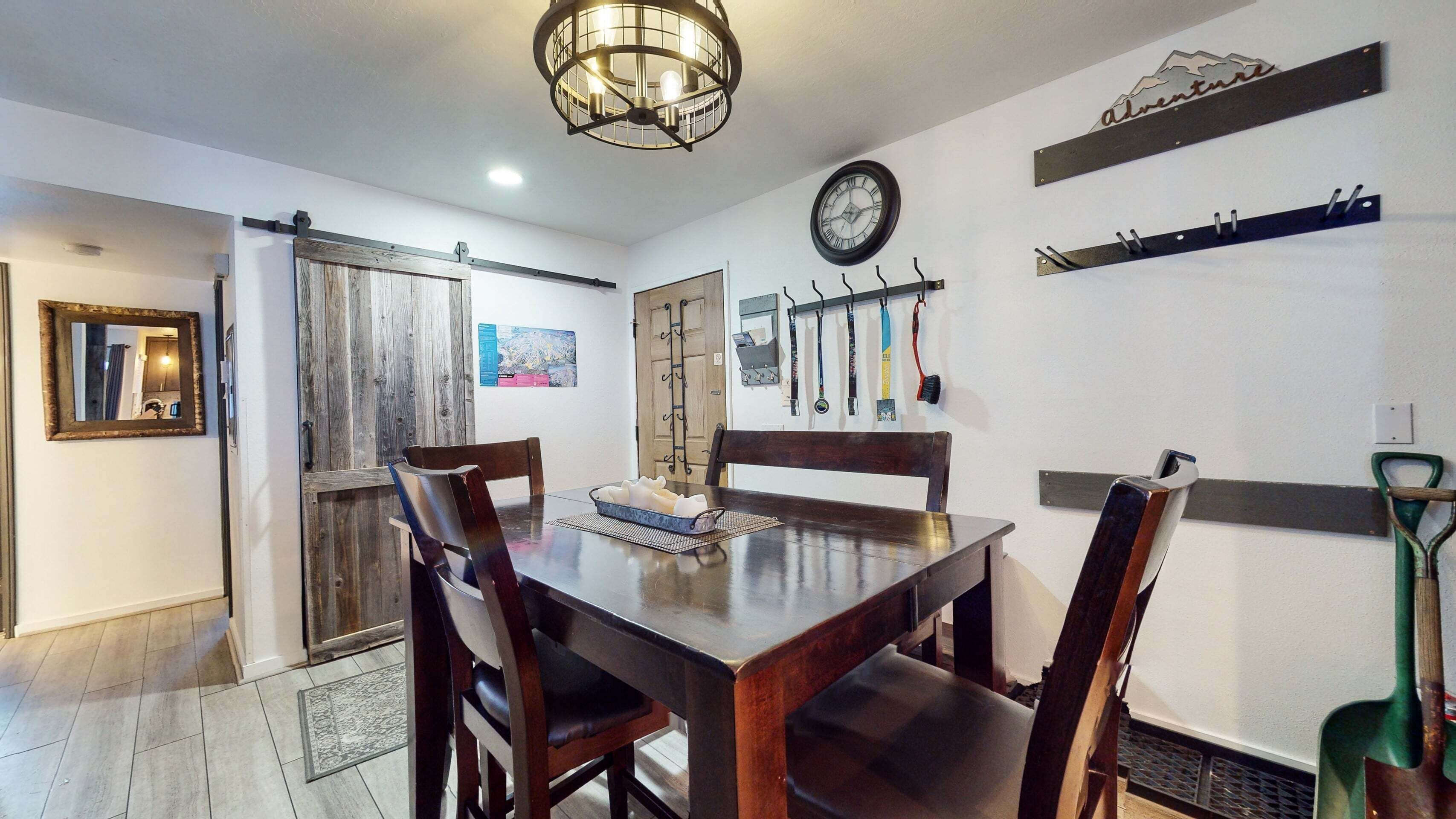 A dining area with a wooden table, chairs, a chandelier, a wall clock, and various tools hanging on hooks.