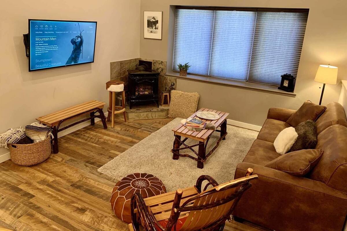 A living room with a fireplace, a television mounted on the wall, a window with blinds, and a brown leather sofa.