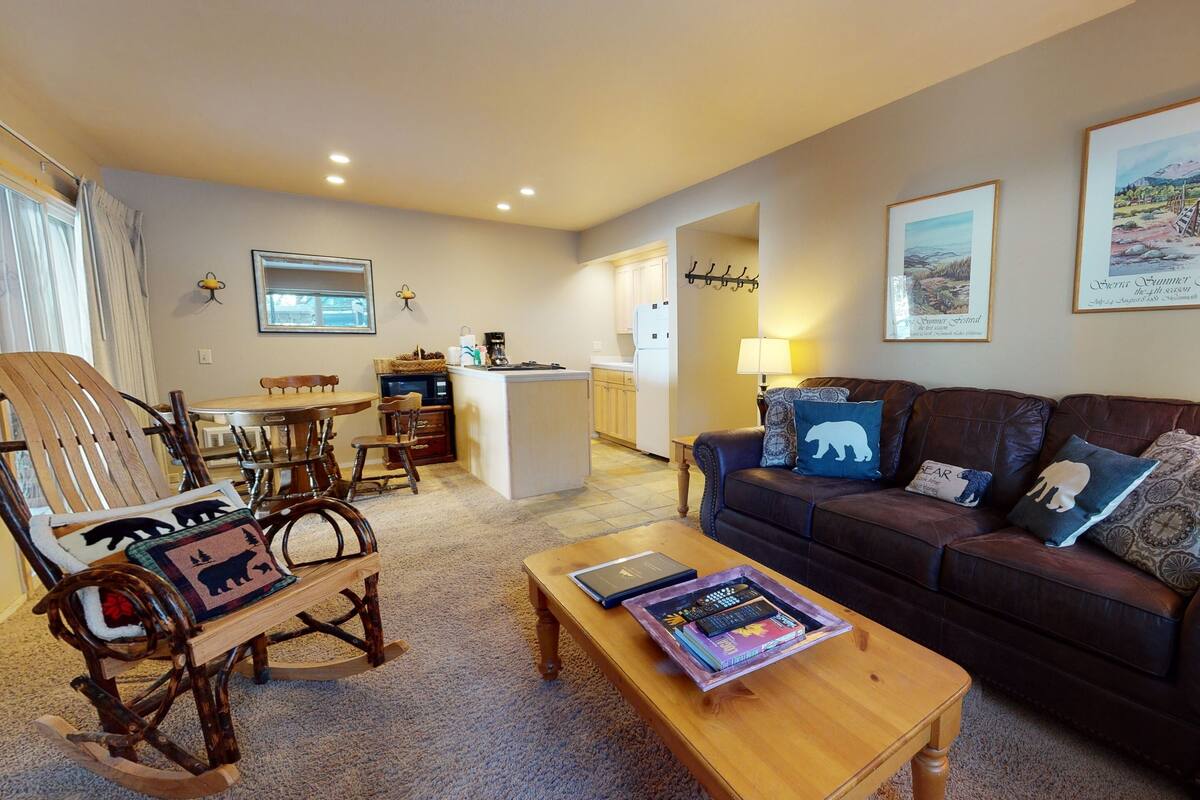 A cozy living room with a wooden rocking chair, a brown leather sofa with bear-themed pillows, a coffee table with books, and a kitchen in the background.