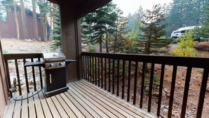 Un terrazza in legno con una griglia, circondata da una ringhiera in metallo e vista su una zona boschiva.