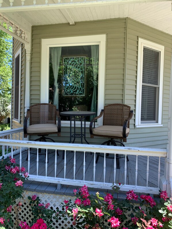 A porch with two wicker chairs and a small table, a decorative window, and a lattice fence with pink flowers.