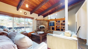 A cozy living room with a stone fireplace, wooden beams, a television, and a bookshelf.