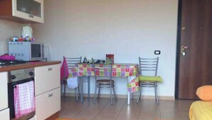 Une petite cuisine équipée d’un four à micro-ondes, d’une cuisinière et d’un four. On y trouve une table à manger avec des chaises et une nappe colorée. Une porte est visible au fond.
