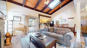 A cozy living room with a sofa, a coffee table, and a fireplace. There is a kitchen area with wooden cabinets and a dining table.