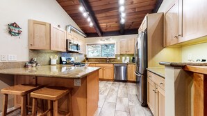 A modern kitchen with wooden cabinets, a granite countertop, stainless steel appliances, and a wooden island with stools. - Mountain Modern, Perfect for Summer, Near The Village, Pool On-site & Hot Tub (Mammoth Lakes)