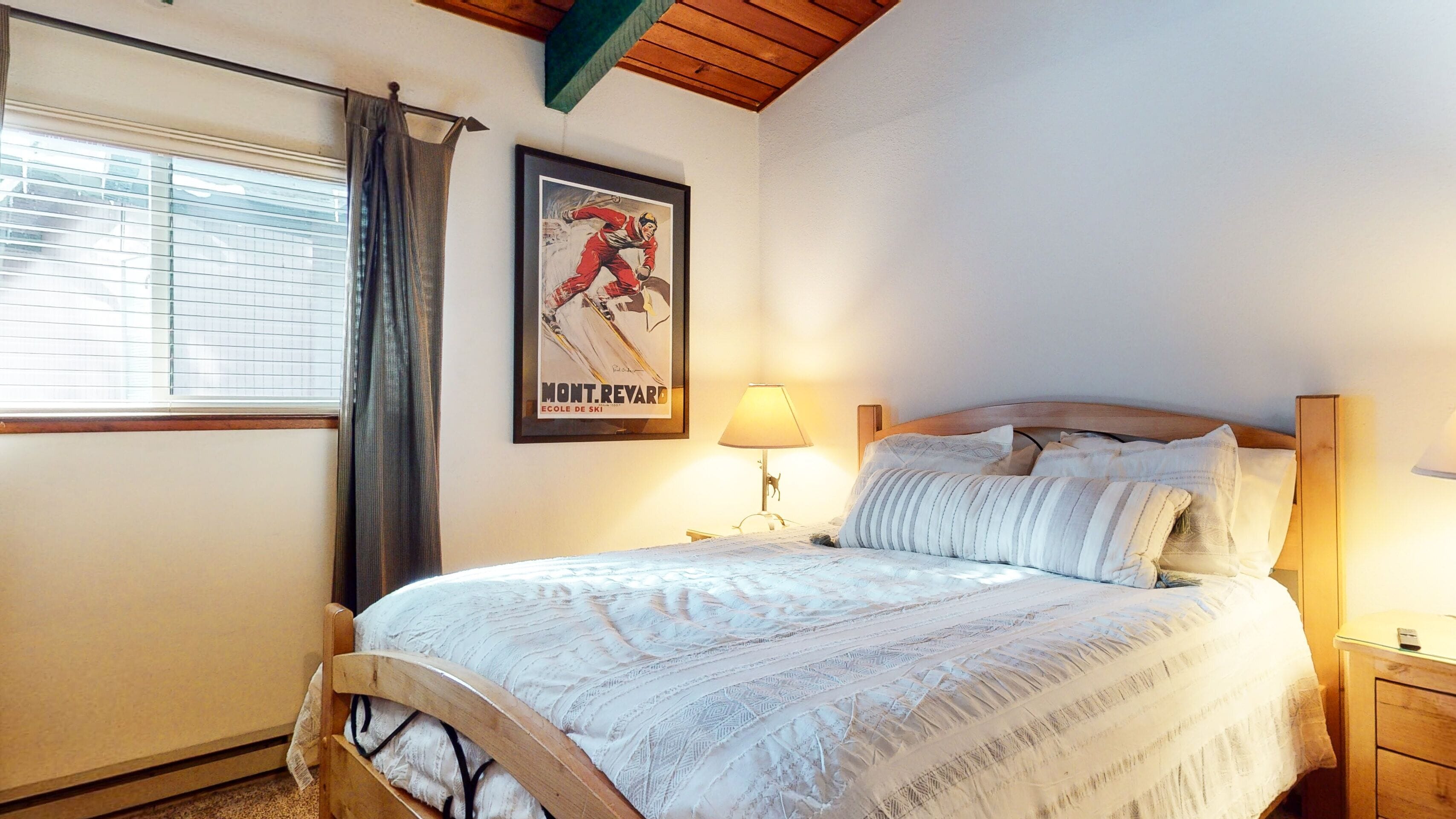 A bedroom with a bed, a nightstand, a lamp, a window with blinds, a curtain, and a poster on the wall.