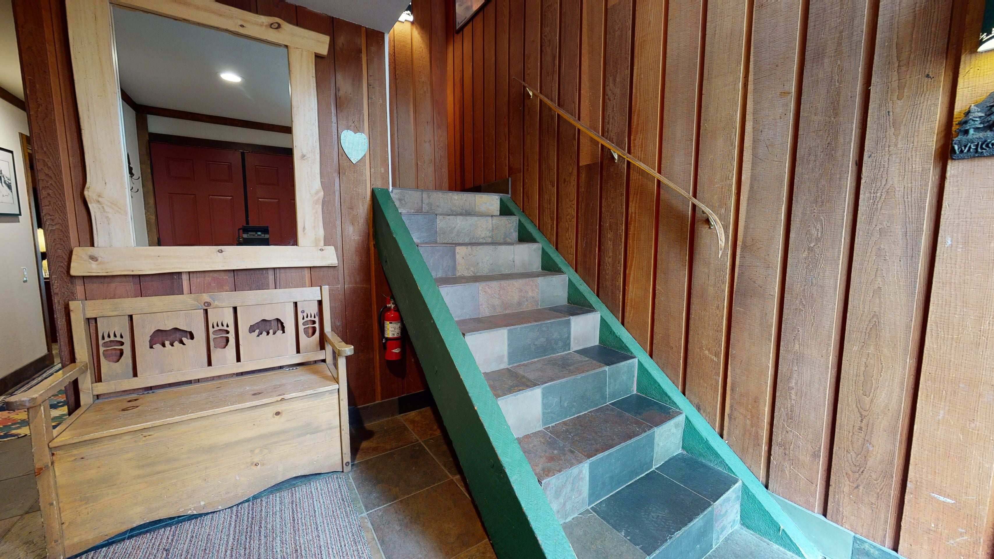 A wooden staircase with a green handrail, a wooden bench with animal carvings, and a fire extinguisher near the stairs.