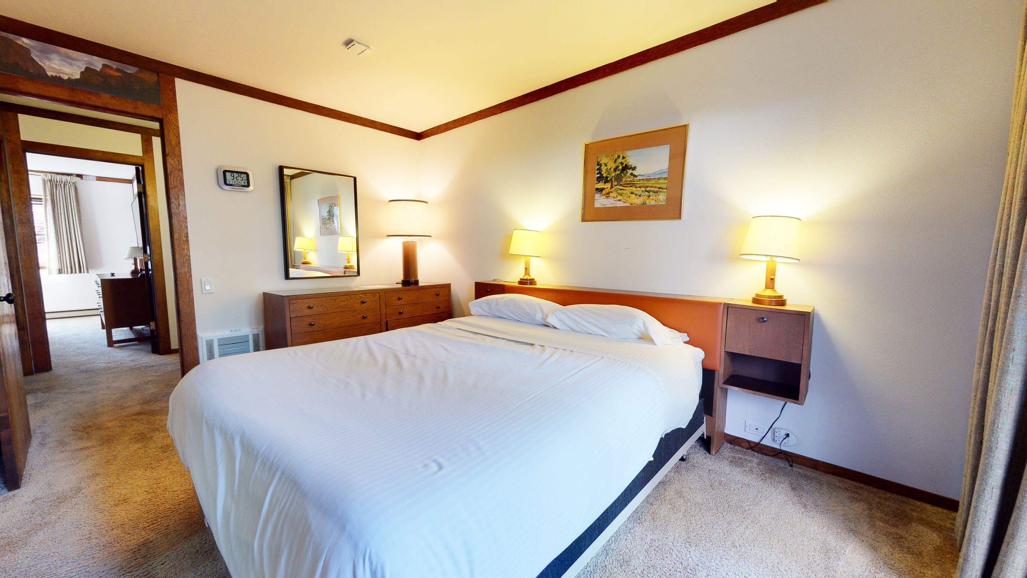 A hotel room with a large bed, two bedside tables with lamps, a dresser, a mirror, and a painting on the wall.