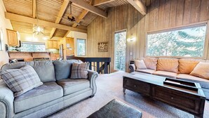 A cozy cabin interior with a wooden ceiling, a sofa, a coffee table, and a large window overlooking trees.