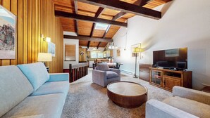 Un salon avec un canapé, des fauteuils, une table basse, un téléviseur et, au fond, une cuisine.