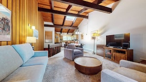 A living room with a sofa, armchairs, a coffee table, a TV, and a kitchen in the background.