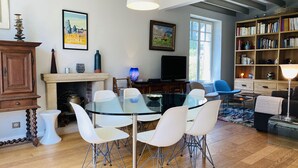 Ett modernt vardagsrum med ett matbord i glas, vita stolar, en öppen spis, en bokhylla och en tv.