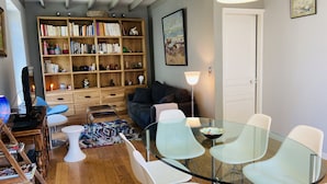 Ett mysigt vardagsrum med ett glasbord omgivet av vita stolar, en bokhylla fylld med böcker och dekorativa föremål, en bekväm soffa och en lampa.