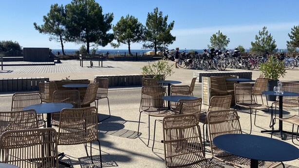 Ett utomhusområde med sittplatser, bestående av flätade stolar och bord, med utsikt över en strand där cyklar står uppställda i bakgrunden.