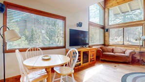Ein gemütliches Zimmer mit einem Holztisch und -stühlen, einem Fernseher und einem Sofa. Große Fenster mit Jalousien sorgen für natürliches Licht.