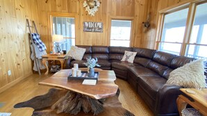 A cozy living room with a wooden interior, a leather sectional sofa, a wooden coffee table, and a welcome sign. - Family and Pet Friendly Mtn Top Cabin, Double Porch, High-Speed WiFi/SmartTVs (Ferguson)