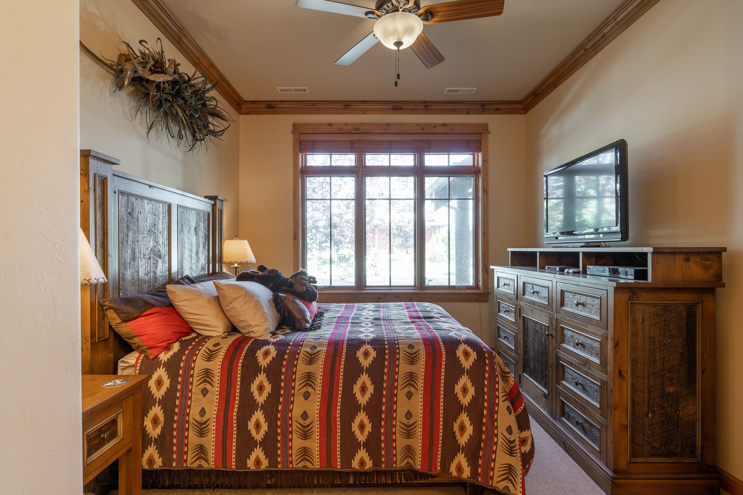 A bedroom with a large bed, wooden headboard, a ceiling fan, a window with blinds, a TV on a wooden stand, and a dresser with drawers.