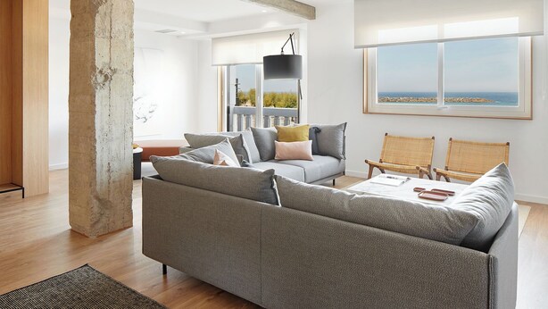 Un salon moderne avec un canapé gris, une table basse et deux fauteuils en osier. Une grande fenêtre offre une vue sur la mer.