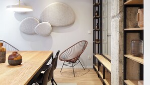 Un espace de salle à manger moderne avec une table en bois, des chaises noires, un luminaire suspendu et un mur en pierre agrémenté d’étagères.