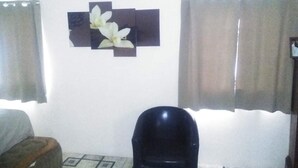 A room with a bed, a black chair, a wall-mounted decoration, and two curtains. - Immaculate 2-bed Apartment in Spanish Town (Spanish Town)
