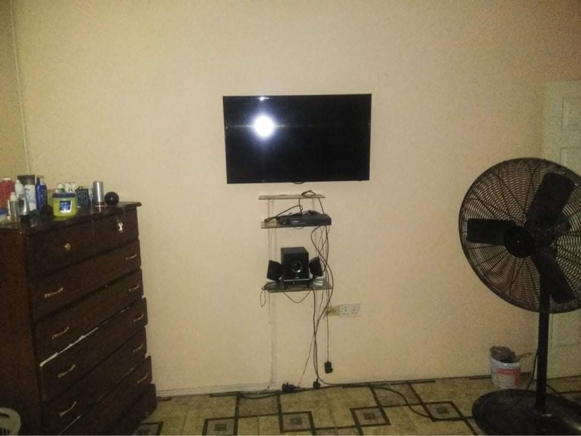 A room with a flat-screen TV mounted on the wall, a small shelf with electronic devices, a fan, and a wooden dresser.