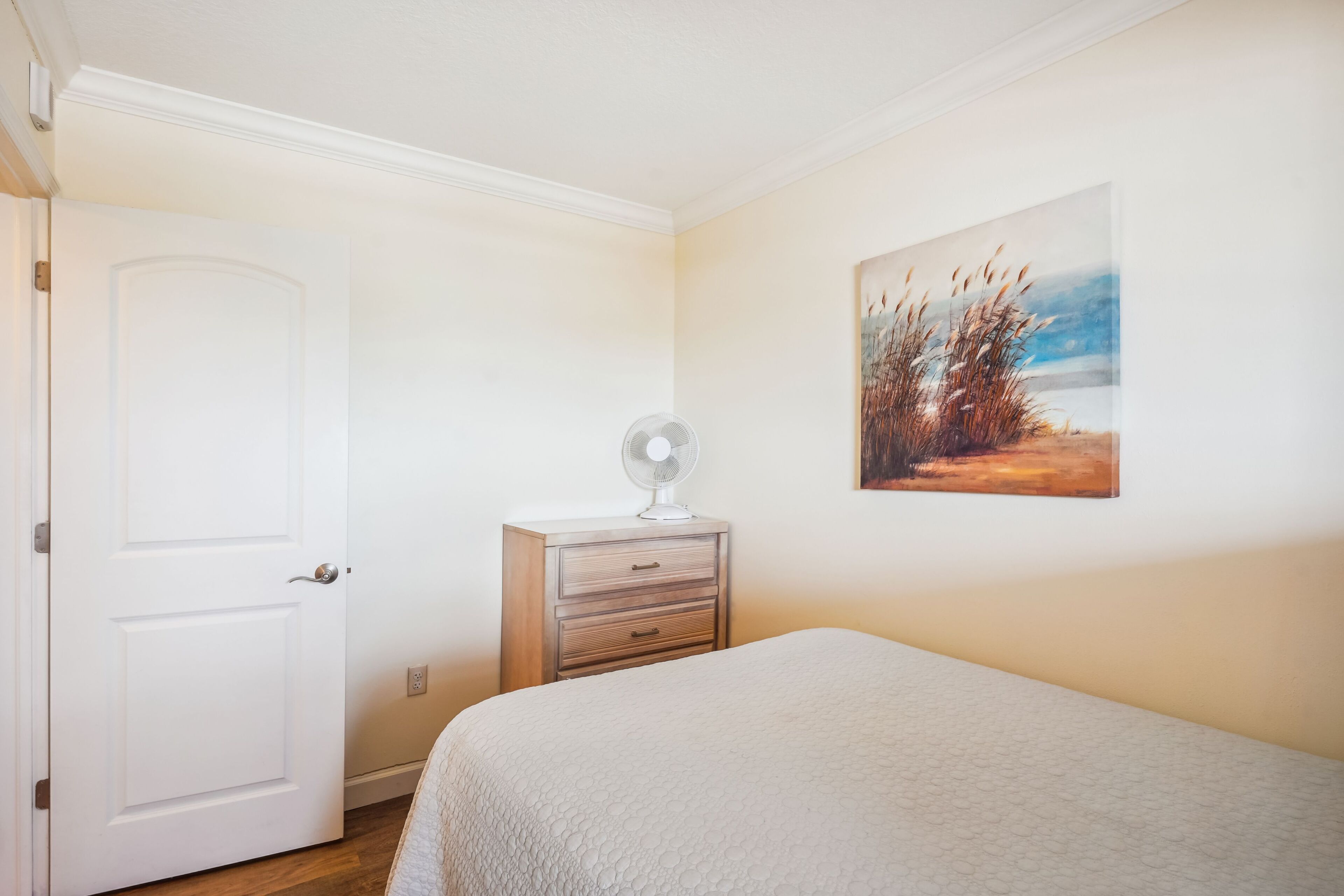 Ein Schlafzimmer mit einer weißen Tür, einem Nachttisch mit Ventilator, einem Bett mit strukturierter Decke und einem Landschaftsbild an der Wand.