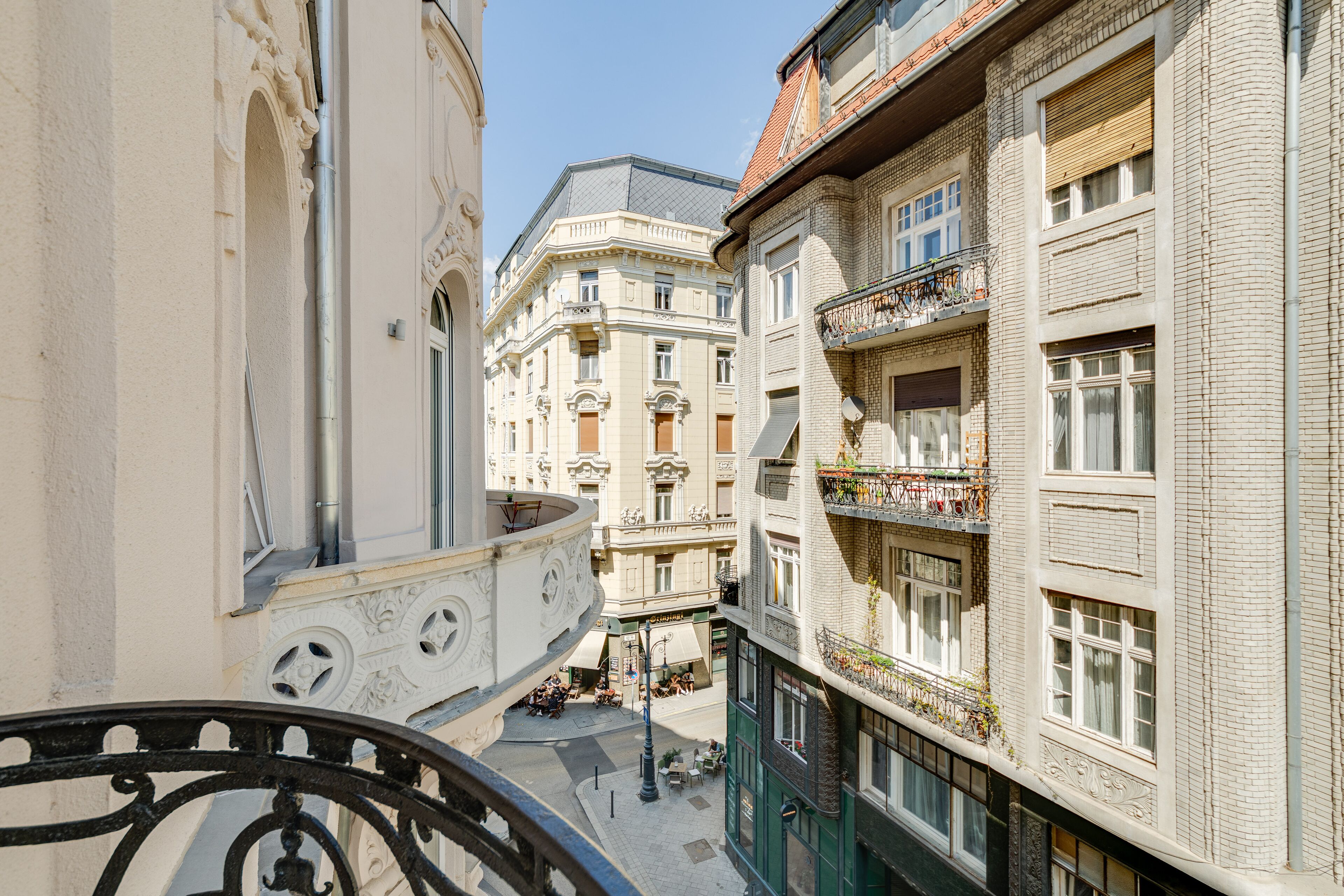 A narrow street with buildings on either side, featuring balconies with plants and a wrought iron railing.