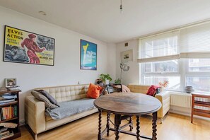 Una sala de estar con un sofá beige, una mesa redonda de madera y dos ventanas con persianas.