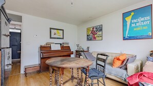 Ein Wohnzimmer mit einem Klavier, einem Esstisch und einer Couch. An den Wänden hängen Poster und eine Tür führt in einen anderen Raum.