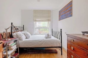 A bedroom with a bed, a bedside table, a dresser, a window with blinds, and a bookshelf.