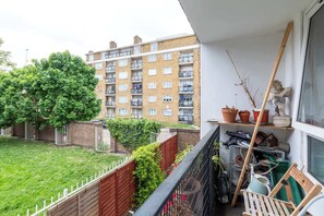 Un balcón con vistas a un edificio de apartamentos de ladrillo, un árbol y una estantería desordenada con diversos objetos.